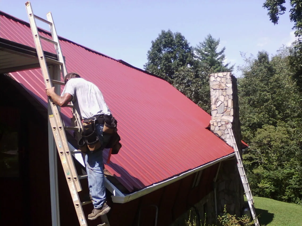 Roofer on a ladder beside a red metal roof and stone chimney — Soffit & Fascia Repair by Roofing Brothers in Saltillo